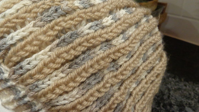 May include: A close-up of a knitted scarf in beige, white, and gray. The scarf is made of a soft, textured yarn and has a braided pattern.