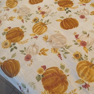 May include: A cream-colored tablecloth featuring a repeating pattern of pumpkins in shades of orange and white, interspersed with sunflowers, leaves, and floral accents. The design evokes a fall or harvest theme.