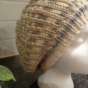 May include: A beige and gray knitted beanie hat with a slouchy fit. The hat is displayed on a white mannequin head.