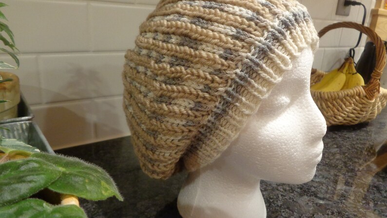 May include: A beige and gray knitted beanie hat with a slouchy fit. The hat is displayed on a white mannequin head.