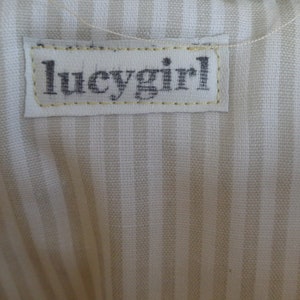 May include: A white fabric with thin, light brown stripes. A white label with black text that reads "lucygirl" is sewn onto the fabric.