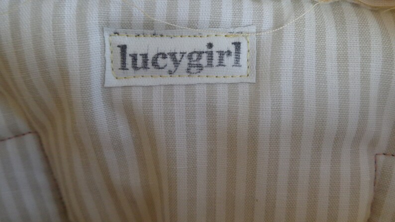 May include: A white fabric with thin, light brown stripes. A white label with black text that reads "lucygirl" is sewn onto the fabric.