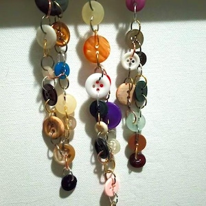May include: Three handmade button necklaces hanging from a wooden dowel. Each necklace features a unique combination of colorful buttons in various sizes and shapes, connected by metal jump rings. The buttons are in shades of black, white, orange, pink, and brown.