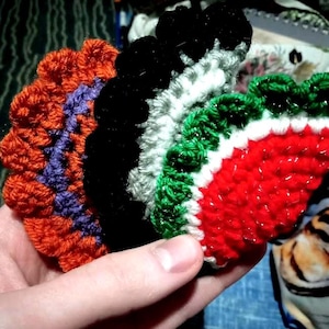 May include: A hand holds a collection of colorful, crocheted items. The items include orange, purple, black, white, green, and red yarn. The crocheted pieces appear to be circular or fan-shaped, with a textured appearance.