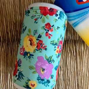 May include: A light blue can cooler with a floral pattern featuring red, yellow, and purple flowers. The cooler is cylindrical and appears to be made of fabric. A bottle of cleaning solution is partially visible in the background.
