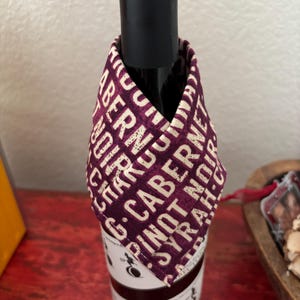 May include: A wine bottle with a purple fabric cover. The fabric has a white print of various wine varietals, including Cabernet, Chardonnay, Pinot Noir, and Syrah.
