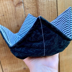 May include: A fabric bowl cozy featuring a black exterior with a repeating pattern of treble clef musical symbols and a contrasting interior lining of thin black and white horizontal stripes, constructed with four distinct corner points.