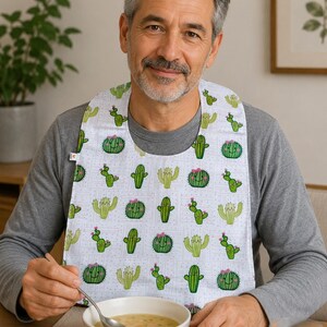 May include: A white adult bib featuring a repeating pattern of green cartoon cacti with smiling faces and small pink bows. The bib has a rounded neckline and covers the chest area, providing protection during meals with a playful, lighthearted design.