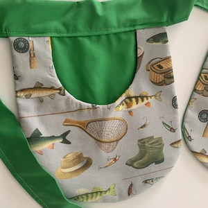 May include: A fishing-themed apron with a green trim and straps. The apron's main fabric is light gray and features illustrations of fish, fishing gear, and boots. The apron is designed with a large pocket.