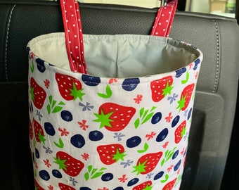 May include: A white fabric car trash bag with a red and white polka dot handle. The bag has a strawberry and blueberry pattern. The bag is hanging from the headrest of a car seat.