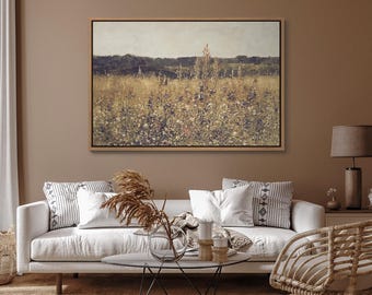 May include: A framed canvas print of a field of wildflowers with a blurred background of trees and a cloudy sky. The print is hanging on a light brown wall above a white couch with throw pillows.