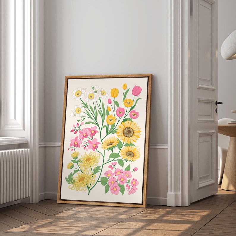 以下が含まれることがあります： 黄色、ピンク、白の色合いの花々が描かれた、額入りの花の芸術プリント。ひまわり、チューリップ、その他の花がクリーム色の背景に配置されています。フレームは明るい茶色です。