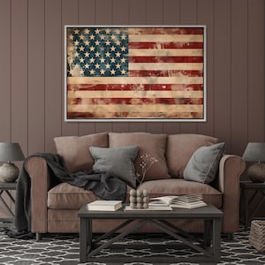 May include: Framed American flag artwork with a distressed, vintage aesthetic. The flag features red and white stripes and a blue field with white stars. The artwork is displayed above a brown sofa with gray pillows.