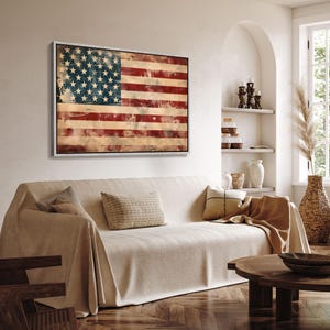 May include: A framed artwork depicting the American flag with a distressed, vintage aesthetic. The flag features red and white stripes and a blue field with white stars. The artwork is displayed above a beige sofa in a living room setting.