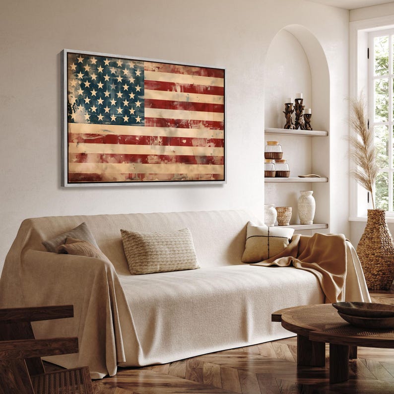 May include: A framed artwork depicting the American flag with a distressed, vintage aesthetic. The flag features red and white stripes and a blue field with white stars. The artwork is displayed above a beige sofa in a living room setting.