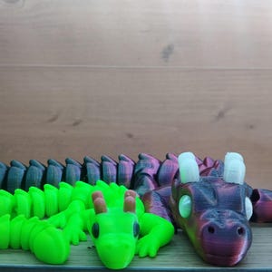 Peut inclure: Deux figurines de dragon imprimées en 3D. Le plus grand dragon est rouge et noir avec des cornes blanches. Le plus petit dragon est vert et noir. Les deux dragons ont un corps segmenté.