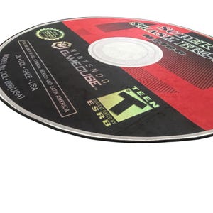 May include: A black and red GameCube game disc with the title "Super Smash Bros. Melee" printed on it. The disc is rated "T" for Teen by the ESRB.