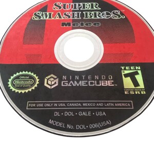 May include: A red and black Nintendo GameCube disc with the text "SUPER SMASH BROS. melee" and "NINTENDO GAMECUBE" printed on it. The disc also has a "TEEN" rating from the ESRB.