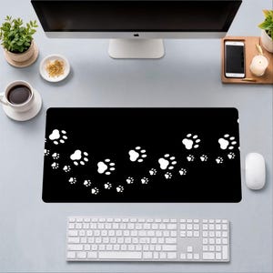 May include: Black rectangular desk mat featuring a repeating pattern of white paw prints. The mat is designed to protect a desk surface and provide a smooth surface for a mouse and keyboard. The design is simple and suitable for pet lovers.