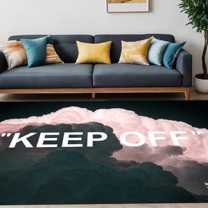 Può includere: Un tappeto nero e bianco con un design a nuvole rosa e il testo "KEEP OFF" in bianco. Il tappeto è in un soggiorno con un divano e una sedia.