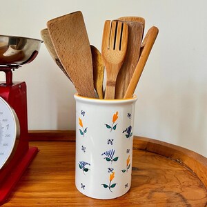 Peut inclure: Un porte-ustensiles en céramique blanche avec un bord jaune et un motif floral en bleu, vert et orange. Le porte-ustensiles est rempli d'ustensiles de cuisine en bois, dont une spatule, une fourchette et une cuillère.
