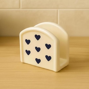 Peut inclure: Porte-serviettes en céramique crème avec un motif de cœurs bleu marine. Le support a un dessus arrondi et une base rectangulaire, conçu pour contenir des serviettes. Le design est simple et élégant, adapté à la décoration de cuisine ou de salle à manger.