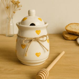 Peut inclure: Pot à miel en céramique blanche avec couvercle, orné de motifs en forme de cœur jaunes. Un nœud en ficelle entoure le pot. Un bâtonnet à miel en bois est posé à côté. Le pot est sur une surface en bois.