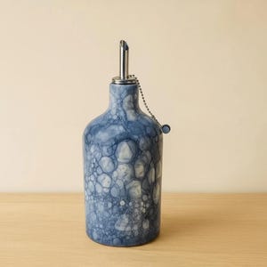 May include: A blue and white ceramic oil bottle with a stainless steel spout and a chain attached to a small stopper. The bottle has a textured, bubble-like pattern. The bottle is sitting on a wooden surface.