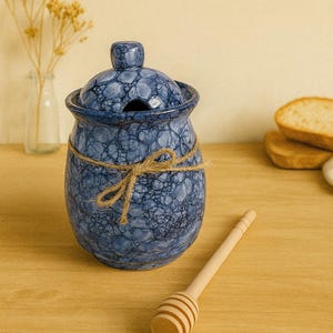 Può includere: Un vasetto di miele in ceramica blu con coperchio, legato con spago, e un cucchiaio per miele in legno. Il vaso ha un motivo strutturato e screziato. Lo sfondo include fette di pane e un vaso con fiori secchi.