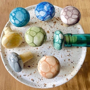 Può includere: Un set di nove tappi per bottiglie in ceramica a forma di fungo. Ogni tappo ha un colore e un motivo di bolle diverso. I tappi sono esposti su un piatto bianco con macchie marroni.