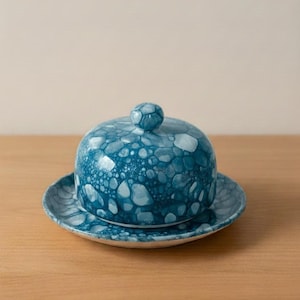 Könnte beinhalten: Ein türkisfarbenes Keramik-Buttergericht mit passendem Deckel und Teller. Der Deckel hat einen kleinen, runden Knauf. Das Gericht und der Deckel haben ein gesprenkeltes Muster in Hellblau und Weiß.