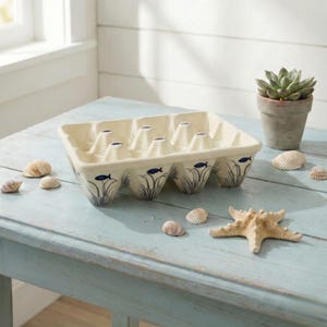 Può includere: Portauova in ceramica color crema con design costiero. Il supporto presenta un motivo di pesci e alghe in blu e grigio. Conchiglie e una stella marina sono su un tavolo azzurro, con una pianta succulenta in vaso sullo sfondo.