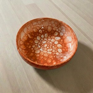 May include: A round, orange bowl with a distinctive pattern of white and orange bubble-like shapes. The bowl has a glossy finish, possibly ceramic or glass. The design gives the bowl a textured appearance.