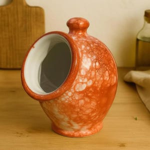 May include: A ceramic salt pig in orange with a white, speckled pattern. It has a round opening and a small knob on top. The interior is white. It is displayed on a wooden surface.