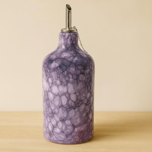 Op de afbeelding: Een decoratieve paarse olieflacon met een roestvrijstalen tuit en ketting. De fles heeft een uniek belachtig patroon in tinten paars en wit. De fles is gemaakt van keramiek en is bedoeld voor gebruik in de keuken.