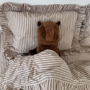 May include: A brown capybara plush toy rests on a bed with striped bedding. The bedding includes a pillow and a comforter, both with ruffled edges. The stripes are a light brown and white color.