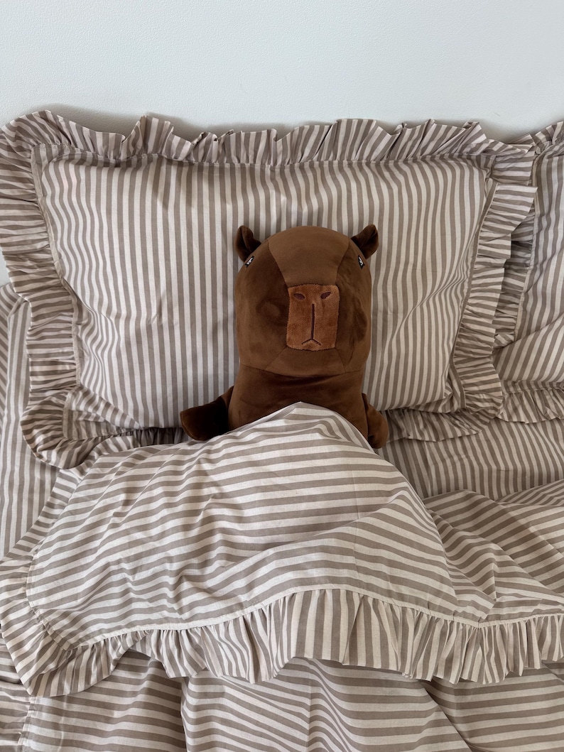 May include: A brown capybara plush toy rests on a bed with striped bedding. The bedding includes a pillow and a comforter, both with ruffled edges. The stripes are a light brown and white color.
