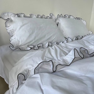 Pode incluir: Roupa de cama branca com babados e acabamento preto. A roupa de cama é feita de um tecido macio e confortável e apresenta um design clássico e elegante.