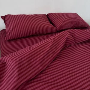 May include: A bedding set featuring a deep burgundy and lighter red striped pattern. The set includes two rectangular pillows and a matching duvet cover, all displaying a consistent vertical stripe design across the fabric surface.