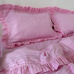May include: Pink and white striped bedding set featuring two ruffled pillowcases and a matching duvet cover. The set has a soft, feminine aesthetic, perfect for a bedroom. The fabric appears to be lightweight and breathable.