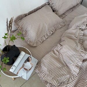 May include: A beige and white striped bedding set with ruffled edges. The set includes a fitted sheet, pillowcases, and a duvet cover. A magazine and decorative items are on a small side table next to the bed.