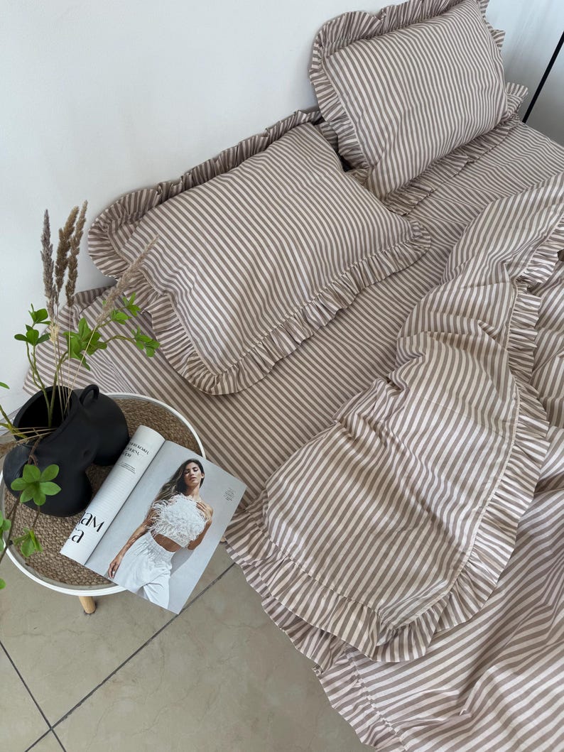 May include: A beige and white striped bedding set with ruffled edges. The set includes a fitted sheet, pillowcases, and a duvet cover. A magazine and decorative items are on a small side table next to the bed.