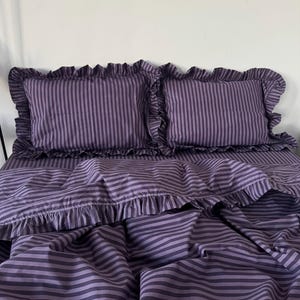 May include: A bedding set featuring two rectangular pillows and a matching duvet cover, all decorated with a dark purple and light purple vertical striped pattern. Each pillow and the duvet cover edge are finished with a ruffled fabric border.