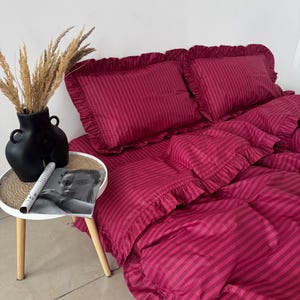 May include: A deep magenta bedding set featuring a subtle vertical stripe pattern and ruffled edges on the pillowcases and duvet cover. The set is displayed on a bed, accompanied by a black vase with dried grasses and a magazine on a side table.