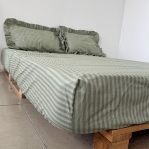 May include: A bed with a light green and white striped bedsheet and two matching pillowcases with ruffled edges. The bed frame is made of wooden pallets. The bedding has a soft, inviting appearance.