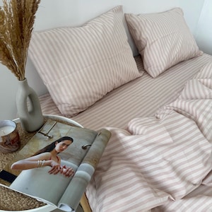 Puede incluir: Ropa de cama a rayas rosa y blancas con una revista en una mesita de noche. La ropa de cama está hecha de tela suave y cómoda y presenta un patrón de rayas sutil.