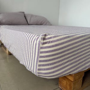 May include: A mattress covered in a fitted sheet featuring a pattern of thin, horizontal purple and white stripes. Two matching striped pillows are positioned at the head of the bed, which rests on a base constructed from wooden pallets.