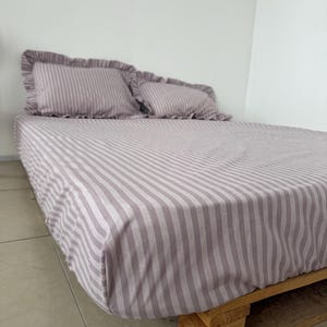 May include: A bed with a striped, mauve-colored fitted sheet and two matching pillowcases with ruffled edges. The bed frame is made of wood. The bedding has a classic, simple design.