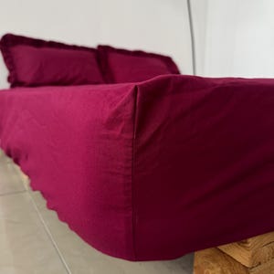 Pode incluir: Um conjunto de cama bordô com lençol com elástico e duas fronhas com bordas com babados. A cama está feita. A cama é sustentada por uma base de madeira.