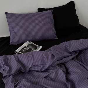 Puede incluir: Una cama con una sábana negra, una almohada a rayas moradas y una funda nórdica a rayas moradas. Una almohada negra está detrás de la almohada morada. Una revista abierta está en la cama.
